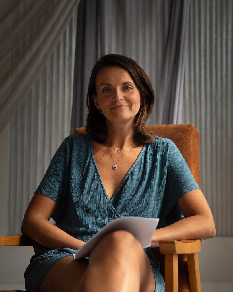 Anna Teresa Kopacz, seated in a wooden chair in a teal dress, papers on her knee, looking at the camera.