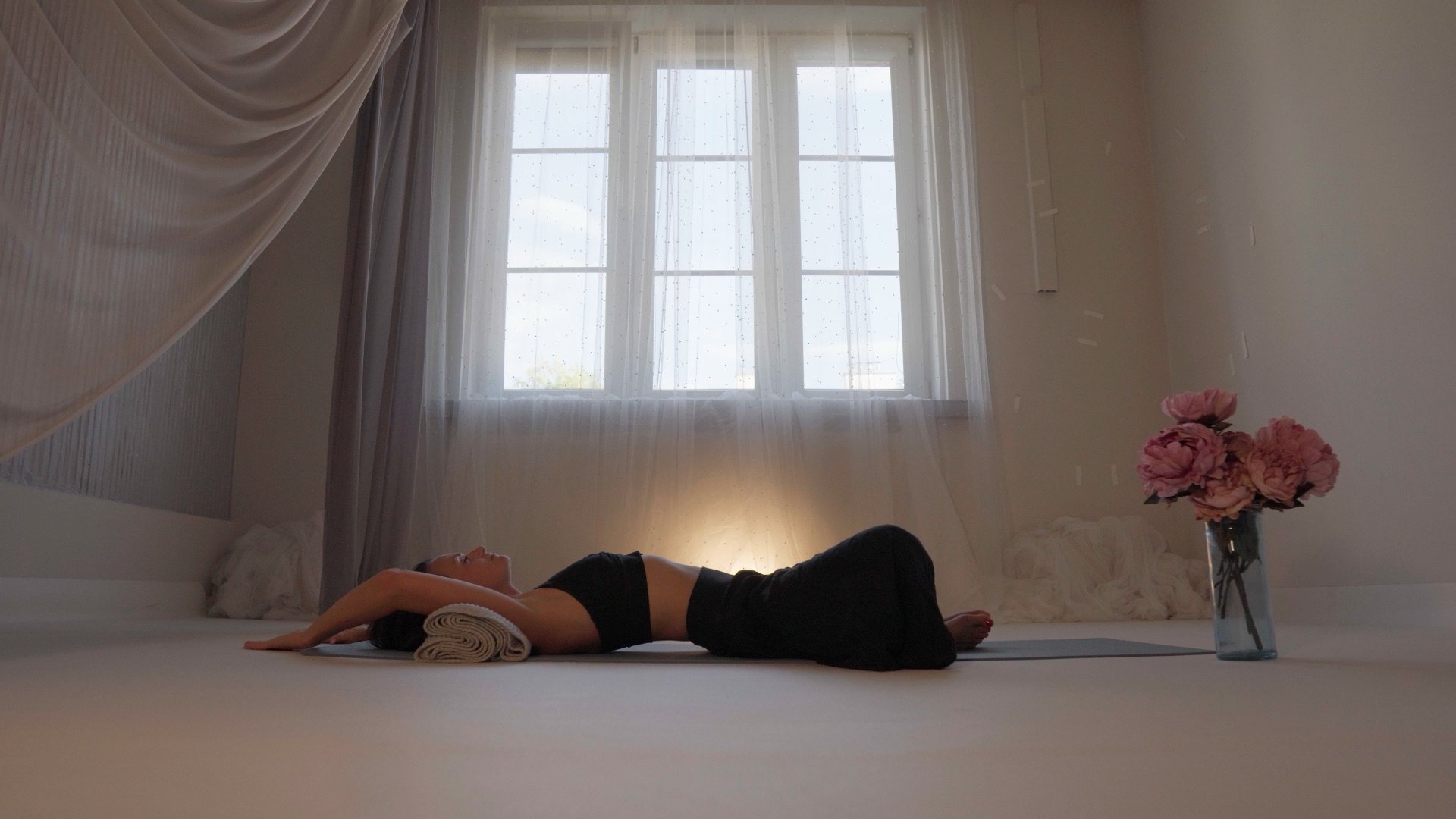 A woman lies on her back on the floor of a light room, tall curtained windows above her, peonies beside her.