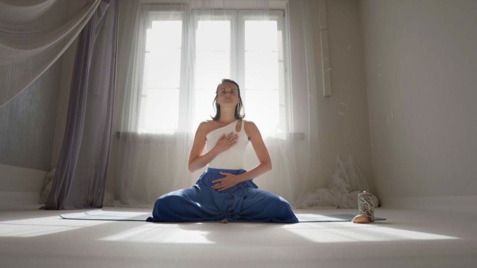A woman sits on the floor in the window light, one hand on her chest, one on her belly.