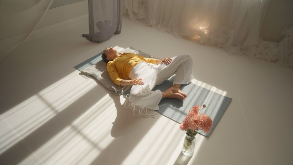 Seen from above, a woman lies on a mat in a light room, sun falling across the floor in long shafts, flowers beside her.